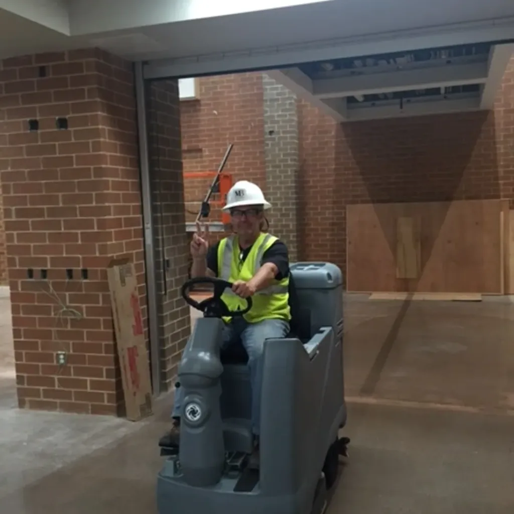 image of a man riding a concrete polishing machine he has a hardhat on and blue jeans hes smiling and holding up a peace sign, he looks very happy