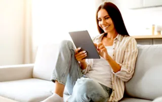 an image of the same Middle Aged Latino Woman Smiling looking at a notepad while she is writing down design ideas for her home's basement, garage, and patio areas, she look very happy sitting on a couch in blue jeans and a loose fitting white tank top and a lite brown and white stripped button up shirt